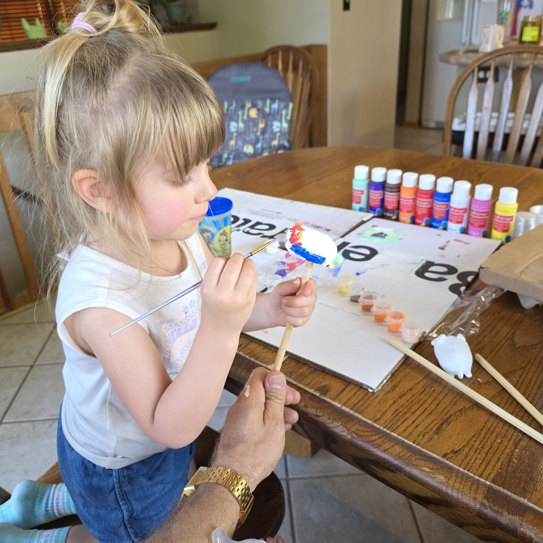 A small child at the kitchen table, brush in hand, painting a bright-white mini figurine on a stick, with a row of craft paints beside her